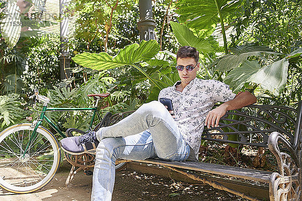 Mann mit Fahrrad  der auf einer Parkbank sitzt und sein Smartphone benutzt