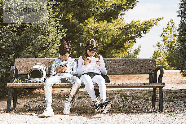 Zwei Kinder in Astronauten- und Superheldenkostümen  die ihr Handy auf der Parkbank benutzen