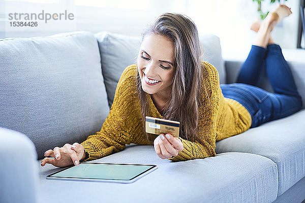 Lächelnde junge Frau benutzt Kreditkarte und Tablet  um online einzukaufen  während sie auf dem Sofa liegt