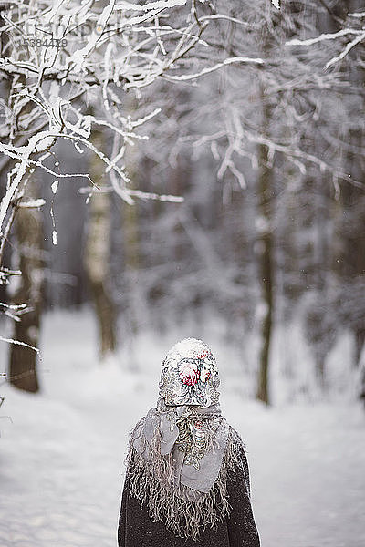 Rückenansicht eines kleinen Mädchens mit Kopftuch  das vor dem Winterwald steht