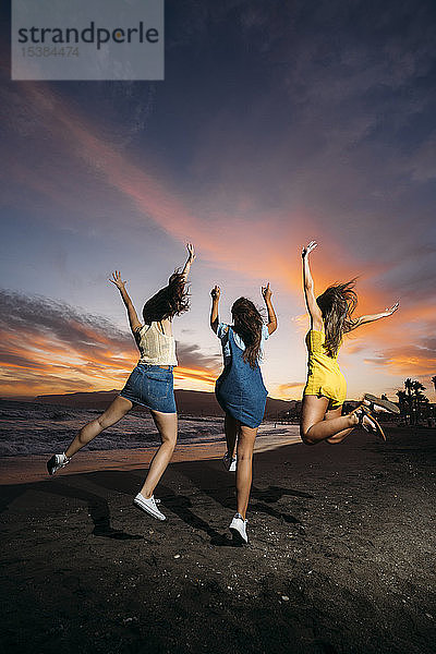 Rückansicht von drei unbekümmerten Freundinnen  die bei Sonnenuntergang an den Strand springen