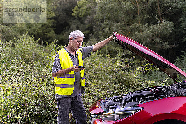 Älterer Mann steht an seinem kaputten Auto  trägt eine Sicherheitsweste und benutzt sein Smartphone