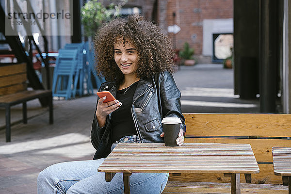 Porträt einer lächelnden Teenagerin  die auf einer Bank sitzt  mit Kaffee zum Mitnehmen und Handy