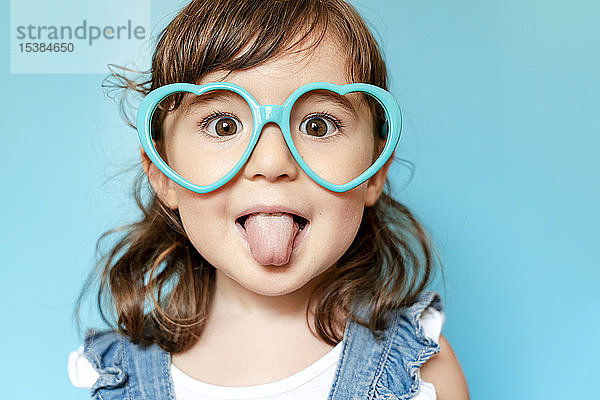 Bildnis eines süßen kleinen Mädchens mit herausgestreckter Zunge und herzförmiger Brille auf blauem Hintergrund