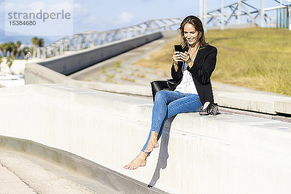 Lächelnde Geschäftsfrau sitzt mit Smartphone und Kopfhörern an der Wand