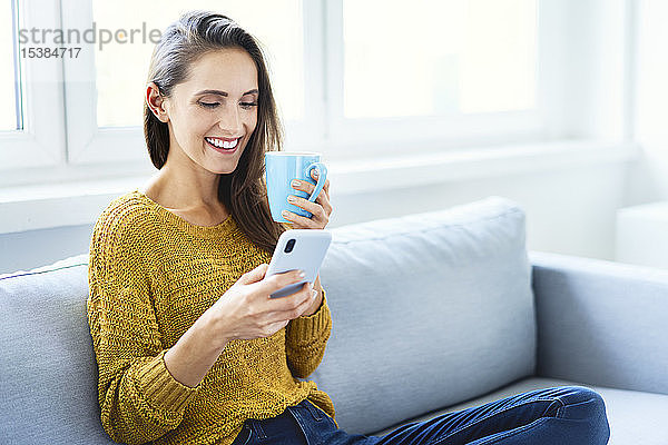 Junge Frau benutzt Smartphone  trinkt Kaffee und sitzt auf dem Sofa