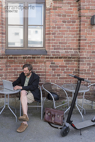 Mann mit Elektroroller sitzt mit Laptop im Straßencafé