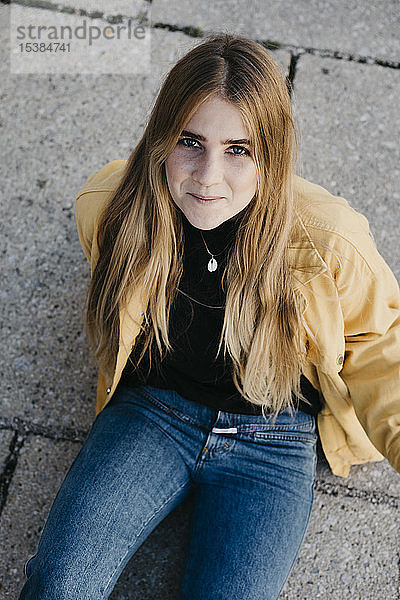 Porträt einer lächelnden jungen Frau in gelber Jacke  die auf dem Boden sitzt