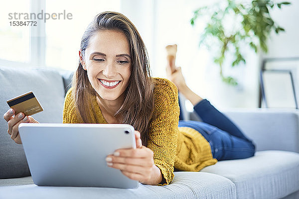 Fröhliche junge Frau benutzt Kreditkarte und Tablet  um von zu Hause aus online einzukaufen