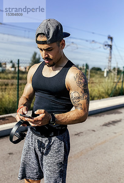 Sportlicher junger Mann mit Smartphone und Kopfhörern auf der Straße stehend