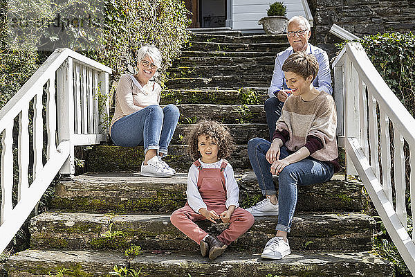 Älteres Ehepaar mit erwachsener Tochter und Enkel sitzen auf einer Treppe im Garten ihres Hauses