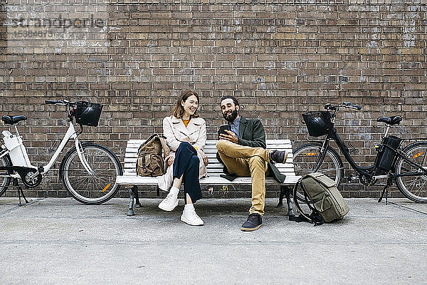 Glückliches Paar sitzt auf einer Bank neben E-Bikes und teilt sein Handy