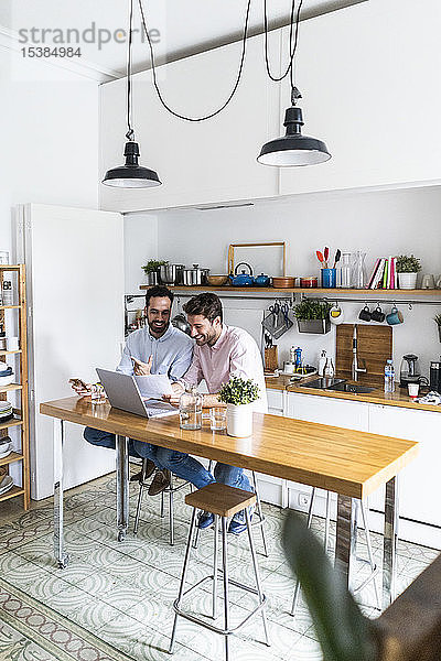 Zwei junge Männer arbeiten gemeinsam in der Küche  benutzen einen Laptop und diskutieren über Dokumente