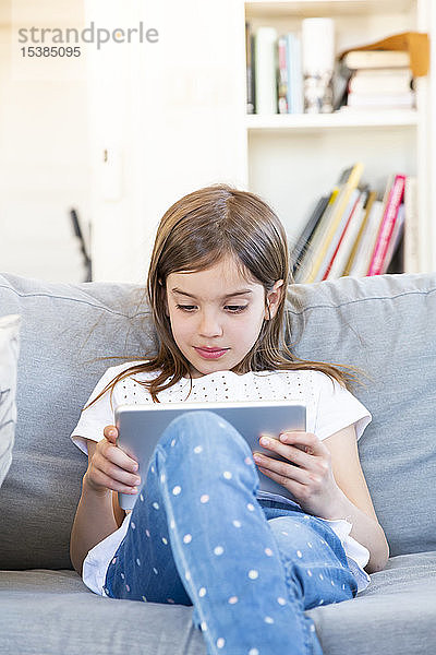 Porträt eines kleinen Mädchens  das zu Hause auf der Couch sitzt und ein digitales Tablet benutzt