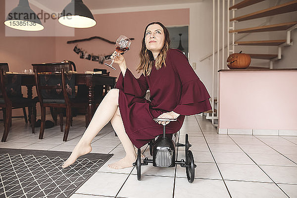 Elegante Frau mit einem Glas Wein  die zu Hause auf einem Tretauto sitzt