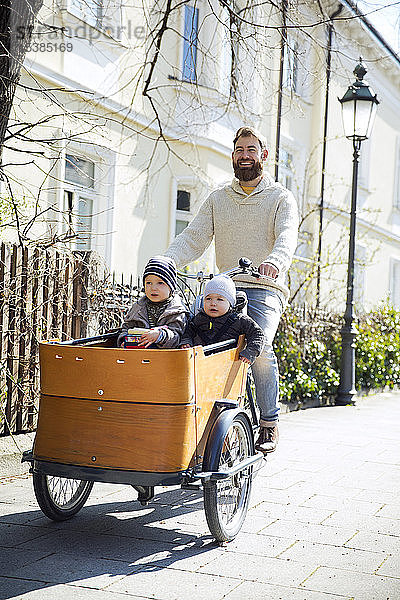 Glücklicher Vater mit zwei Kindern auf dem Lastenfahrrad in der Stadt