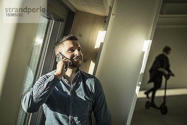 Junger Geschäftsmann spricht am Telefon  während ein Kollege im Hintergrund Kickboard oin