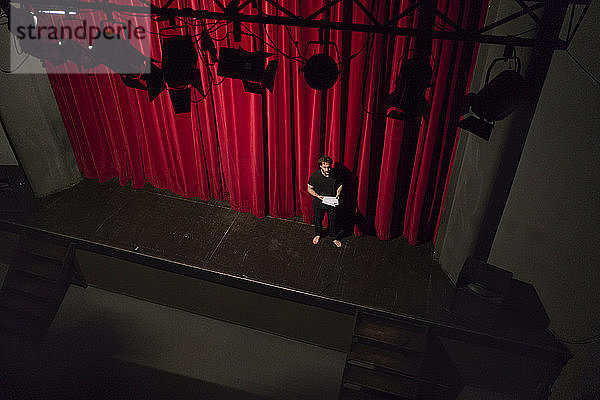 Barfüssiger Schauspieler mit Drehbuch steht auf Theaterbühne vor rotem Vorhang