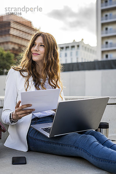 Junge Geschäftsfrau sitzt auf einer Treppe in der Stadt  arbeitet mit einem Laptop  hält ein Dokument