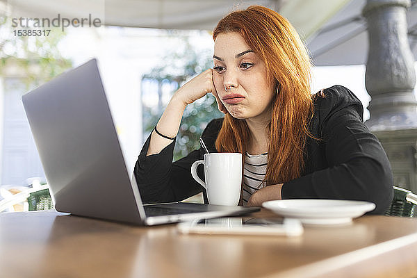 Porträt einer rothaarigen jungen Frau im Straßencafé  die auf ihren Laptop schaut