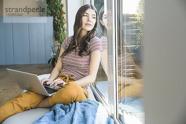Junge Frau sitzt zu Hause mit Laptop am Fenster
