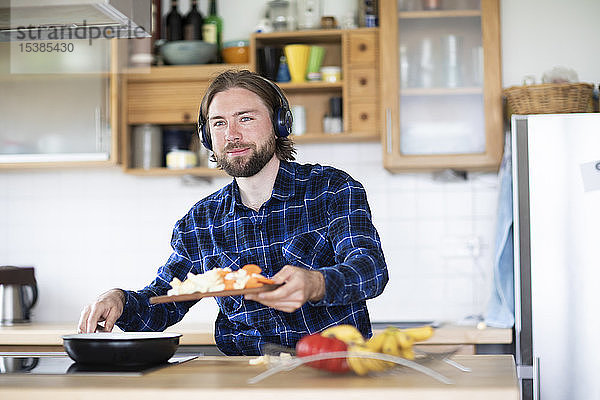 Junger Mann mit Bart und kariertem Hemd und Kopfhörern  der in der Küche Gemüse kocht
