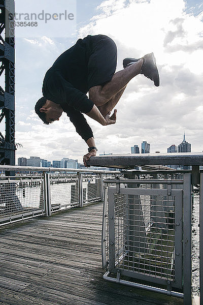 USA  New York  Brooklyn  junger Mann macht Parkour-Handstand auf dem Geländer des Piers vor der Skyline von Manhattan