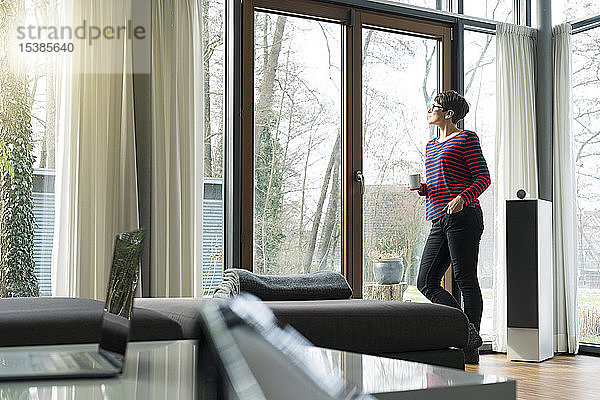 Frau mit Kaffeebecher steht im Wohnzimmer und schaut aus der Terrassentür