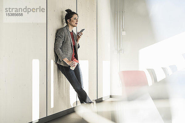 Geschäftsfrau lehnt mit ihrem Handy im Büro gegen eine Wand