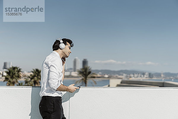 Spanien  Barcelona  Geschäftsmann hört Musik mit Kopfhörern und Smartphone auf der Dachterrasse