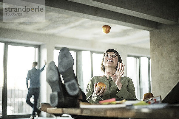 Frau  die im Büro mit Äpfeln jongliert und mit den Füßen auf dem Schreibtisch sitzt