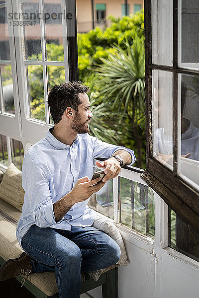 Junger Mann sitzt auf Bank am Fenster und benutzt Smartphone