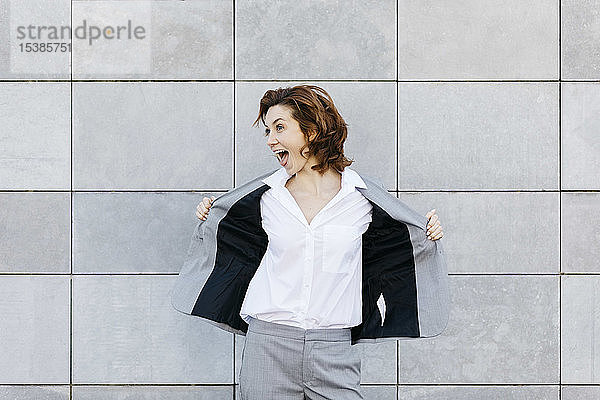 Porträt einer schreienden jungen Geschäftsfrau vor einer Wand mit grauen Kacheln  die ihre Jacke öffnet