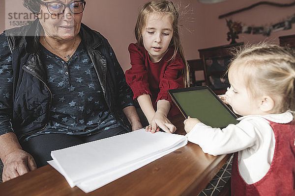Zwei Mädchen und ihre Großmutter spielen im Wohnzimmer mit Papier und Tablette