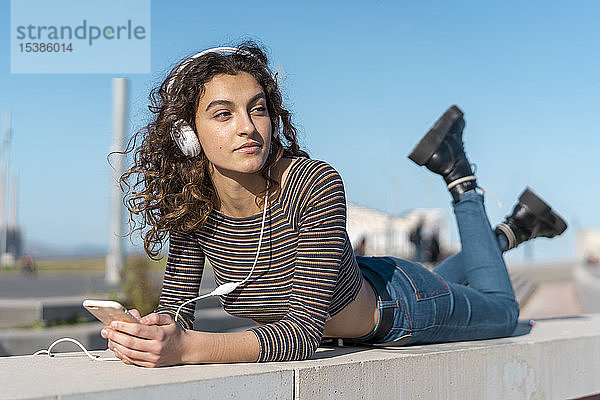 Junge Frau liegt an einer Wand und hört Musik mit Kopfhörern auf ihrem Smartphone