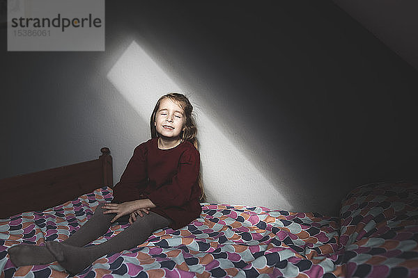 Lächelndes Mädchen sitzt zu Hause mit geschlossenen Augen auf dem Bett