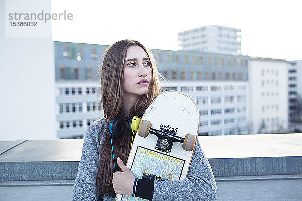 Seriöse junge Frau mit Skateboard und Kopfhörern in der Stadt  die zur Seite schaut