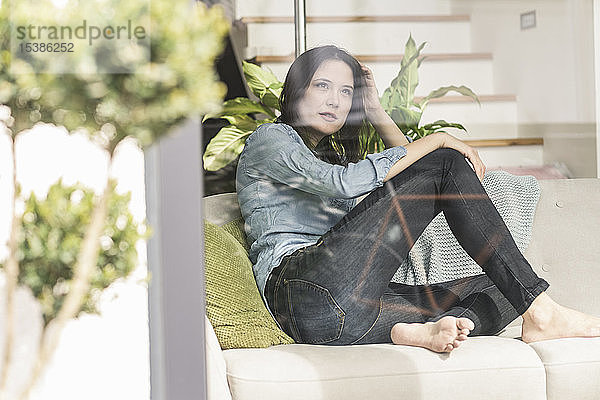 Nachdenkliche Frau sitzt zu Hause auf dem Sofa hinter einer Fensterscheibe