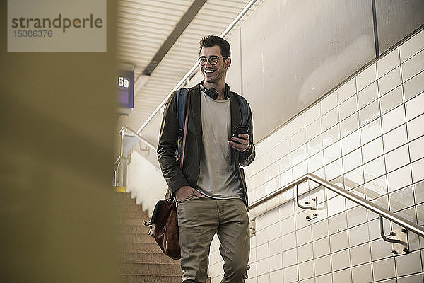 Lächelnder junger Mann mit Handy geht am Bahnhof die Treppe hinunter