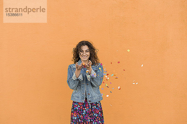 Porträt einer lächelnden jungen Frau  die vor einer orangefarbenen Wand Konfetti in die Luft wirft