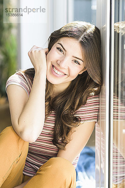 Porträt einer lächelnden jungen Frau  die zu Hause am Fenster sitzt