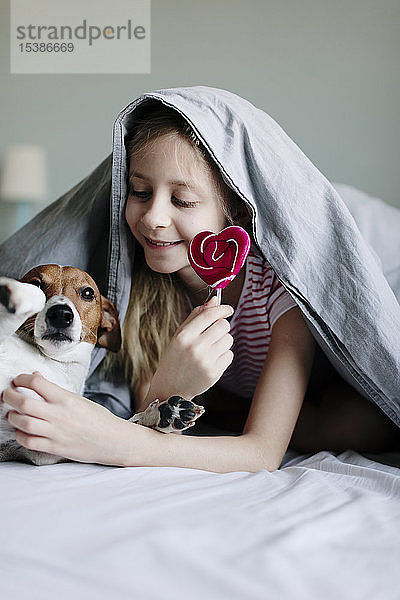 Lächelndes Mädchen liegt auf dem Bett mit einem Lolli  der ihren Hund kitzelt