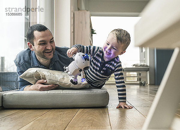 Aufgeregter Vater und Sohn liegen zu Hause auf einer Matratze und schauen einem Spielzeugroboter zu