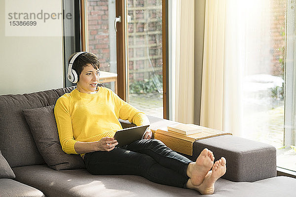 Porträt einer Frau mit digitalem Tablett  die zu Hause auf der Couch sitzt und mit Kopfhörern Musik hört