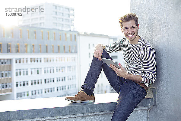 Lächelnder junger Mann sitzt mit Tablette an der Wand