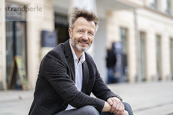 Porträt eines lächelnden reifen Geschäftsmannes mit grauem Bart  der auf Stufen im Freien sitzt