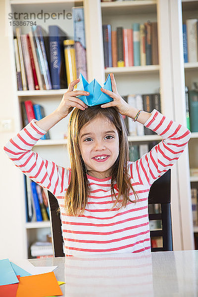 Porträt eines lächelnden Mädchens  das sich zu Hause einen selbst gebastelten Papierhut aufsetzt