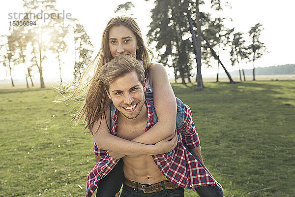 Porträt eines glücklichen jungen Mannes  der seine Freundin huckepack auf der Wiese trägt