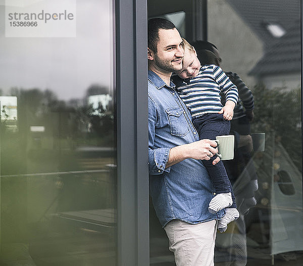 Vater mit Tasse Kaffee trägt Sohn an Terrassentür zu Hause
