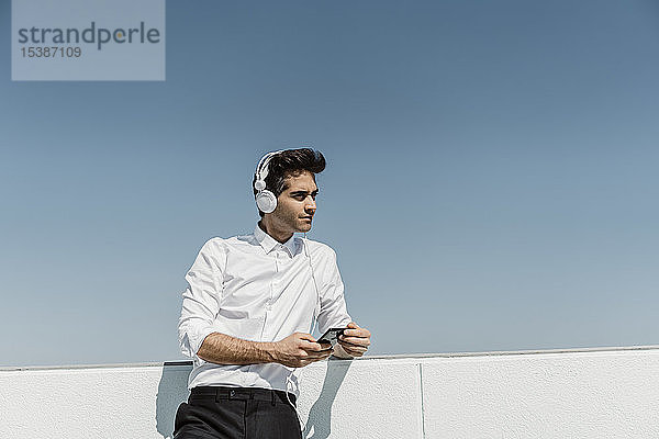 Porträt eines Geschäftsmannes  der auf der Dachterrasse mit Kopfhörern und Smartphone Musik hört